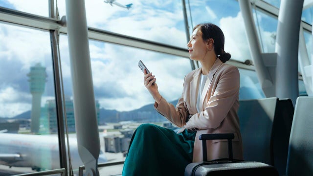 Confident young Asian businesswoman with suitcase looking ahead while using smartphone waiting for her flight at airport terminal. Business traveller. Woman in business. Business lifestyle and technology. Travel and vacation concept 