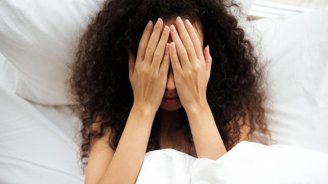 A dark-skinned woman lies in bed and covers her face with her hands. Black girl in bed. Heavy morning. Reluctance to wake up and go to work. Bad dream. Sleeplessness. 