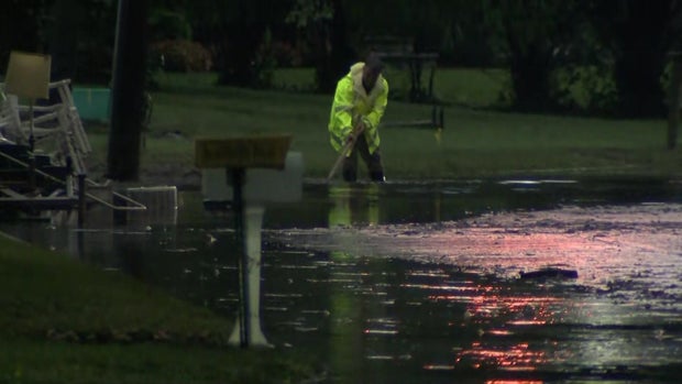 An individual in a neon yellow rain coat tries to clear a drain on a flooded road 