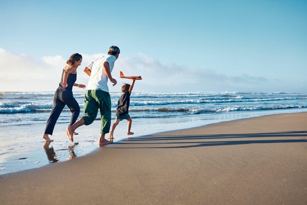Parents running behind a boy running straight ahead with a model airplane held high in one hand. 