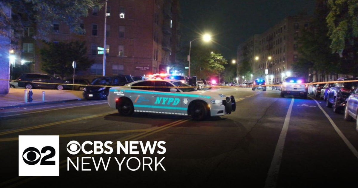 Nighttime city street in the Bronx blocked by an NYPD cruiser with flashing lights; additional police vehicles and orange crime-scene tape cordon off the area in front of apartment buildings.