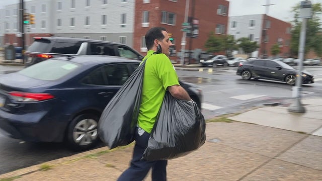 A man wearing a lime green shirt carries two trash bags 
