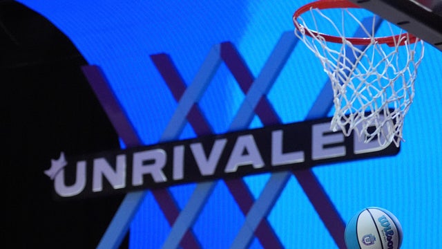 The Unrivaled basketball logo is seen during a game, the net and a basketball are in the foreground 