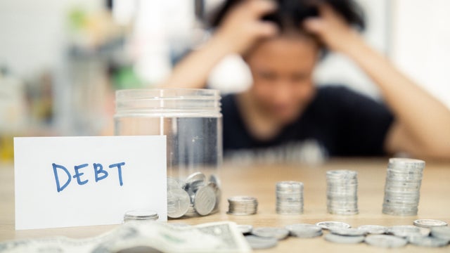 A woman sits at a table, looking distressed, with piled coins and a debt note. This scene expresses the concept of financial burden, economic challenges, and personal reflections on money management. 