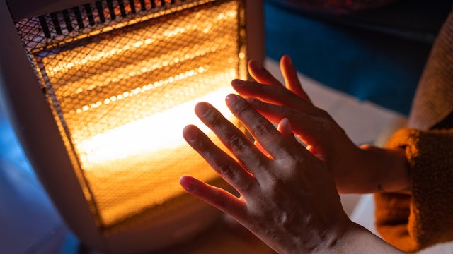 A woman warms her hands on an electric heater at home in the 