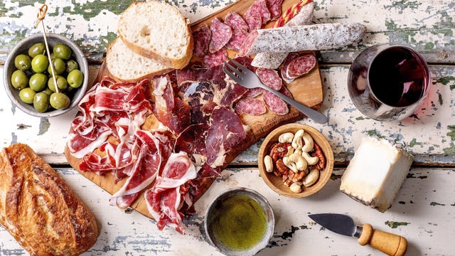Snack Board with Traditional Spanish Jamon Iberico. Sliced Fuet Sausage. Cheese. Green Olives. White Bread. Nuts. Olive Oil. Glass of Red Wine. Light Rustic Wooden Background. Top View 
