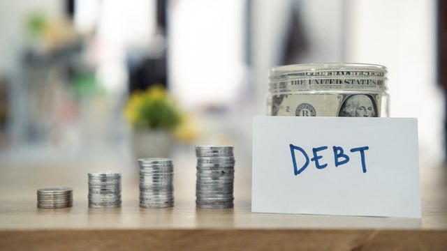 Close-up of stacked coins beside a note labeled debt with a jar filled with cash in the background, symbolizing financial burdens, savings, or economic planning issues. 