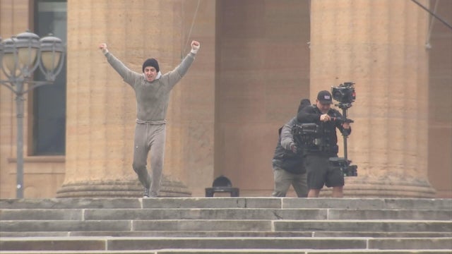 An actor celebrates after running to the top of the Art Museum steps, a cameraman is recording from behind him 
