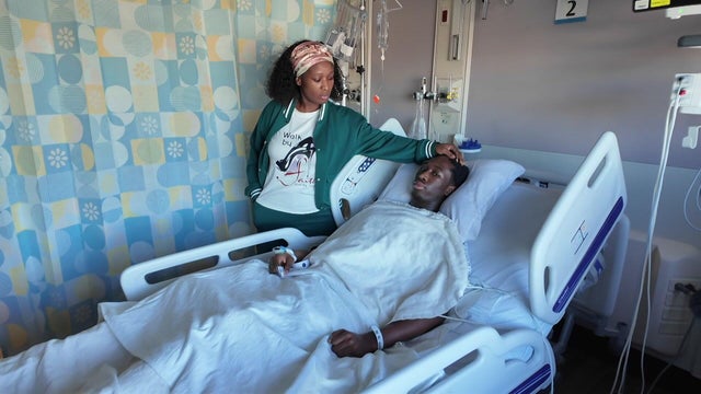A 16-year-old lays in a hospital bed as his mother stands by his bedside 