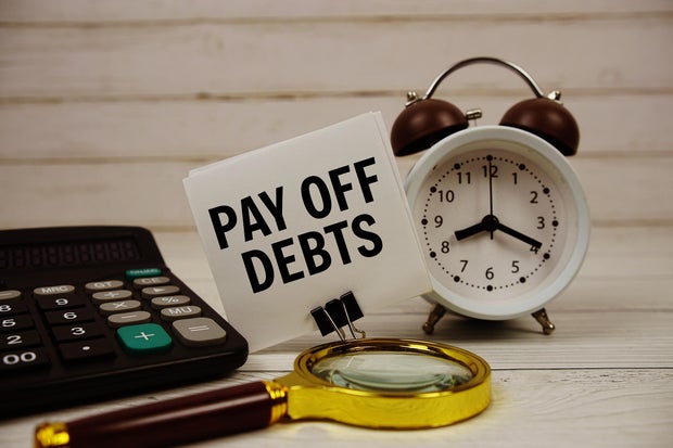Pay Off Debts text on paper card with alarm clock, calculator and magnifying glass on wooden background 