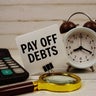 Pay Off Debts text on paper card with alarm clock, calculator and magnifying glass on wooden background 