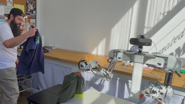 A clothes-folding robot picks up a shirt from a table as a person places more laundry on it. 