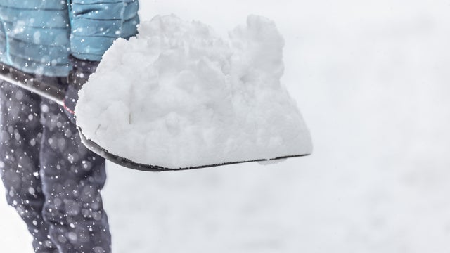 An adult man holds a shovel with a large pile of freshly fallen snow in the middle of a snowfall. 