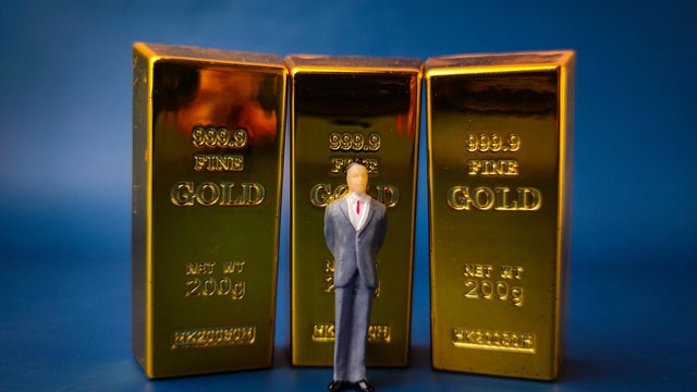 This image depicts a miniature figure of a businessman standing in front of three gold bars, symbolizing business and financial concepts 