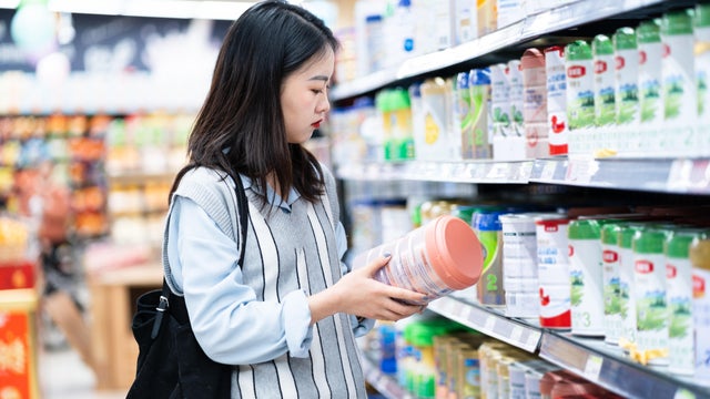 Rear view of young Asian mother with a shopping cart grocery shopping for baby products in a supermarket. She is standing in front of the baby product aisle and have no idea which product to choose from 
