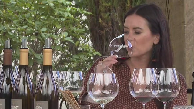A woman holds a wine glass to her mouth as she sits in front a row of glasses and bottles 