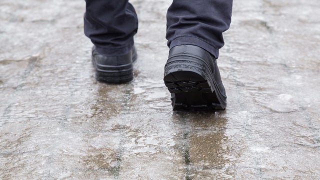 Young man's legs in black leather boots walking on sidewalk in wet, warm winter day. Pavement covered with slippery ice. Closeup. Back view. 