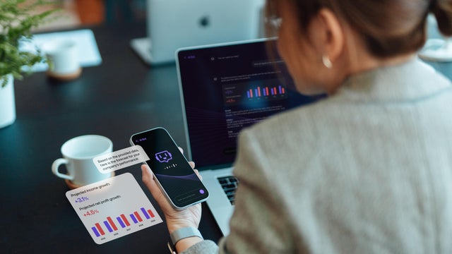 Asian businesswoman using AI chatbot technology on smartphone to assist her while working on laptop in office 