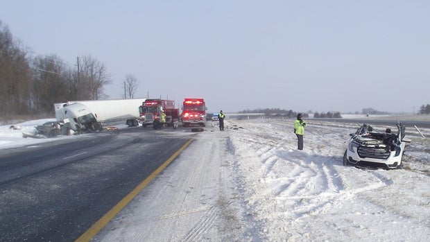 A photo of the December 24, 2022 crash, showing a white SUV with the top ripped off on the right, fire trucks blocking the highway and a crumpled grey pickup truck and a damaged white semi-truck, both in a ditch on the left. 