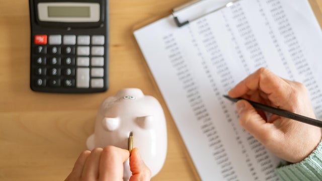 Accountant inserting a coin into piggy bank and calculating expenses with calculator and spreadsheet on a wooden desk, managing home budget and savings 