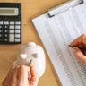 Accountant inserting a coin into piggy bank and calculating expenses with calculator and spreadsheet on a wooden desk, managing home budget and savings 
