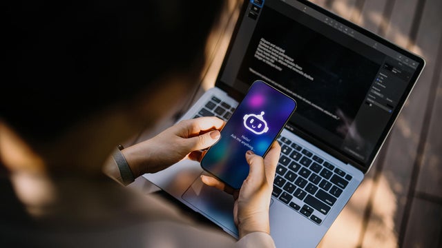 Businesswoman using Chatting AI chatbot virtual assistance app on smartphone to assist her while working on laptop in office park. Using AI technology at work. Online robot system. Concept of VR and AR technologies. People, AI, innovation and technology 