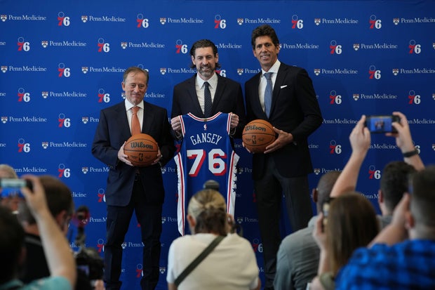 Mike Gansey holds a Sixers jersey with his name, flanked by Josh Harris and Bob Myers, both holding basketballs, all in front of a 76ers-branded backdrop 