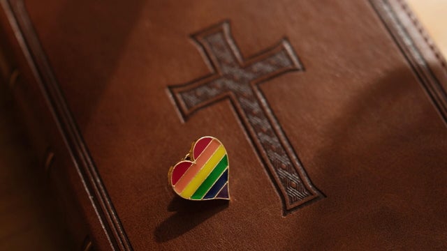 A book with a cross on it, a pin in the shape of a heart colored like a Pride flag is laid on top 