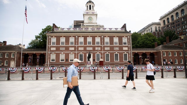 People walk in front of Independence Hall, the fence is decorated with bunting for July Fourth 
