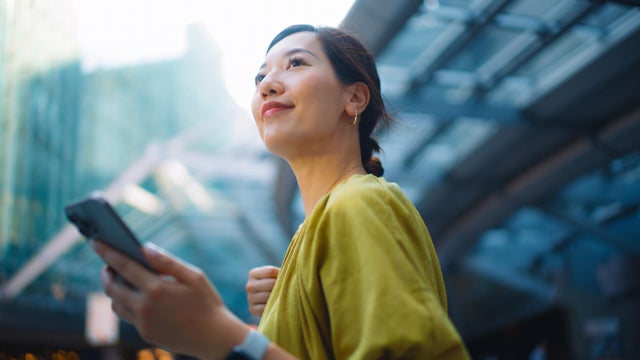 Young Asian businesswoman using online banking with mobile app on smartphone to manage her banking needs in the city, against financial skyscrapers on the go. Managing finance and investment at easy access. Wealth management. Smart banking with technology 