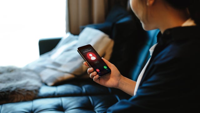 A woman receiving an incoming suspected call from unknown caller on her smartphone and rejecting the call at home. Device screen showing warning sign as detected by network provider. Phone scam. Cyber security and fraud concept 