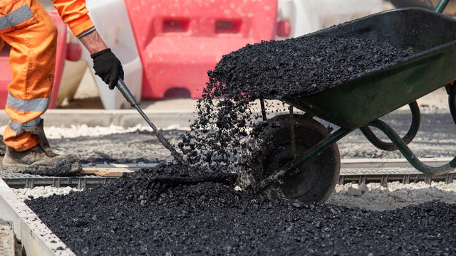 Team of road workers in bright orange uniforms repair road surface with wheelbarrow filled with hot asphalt 